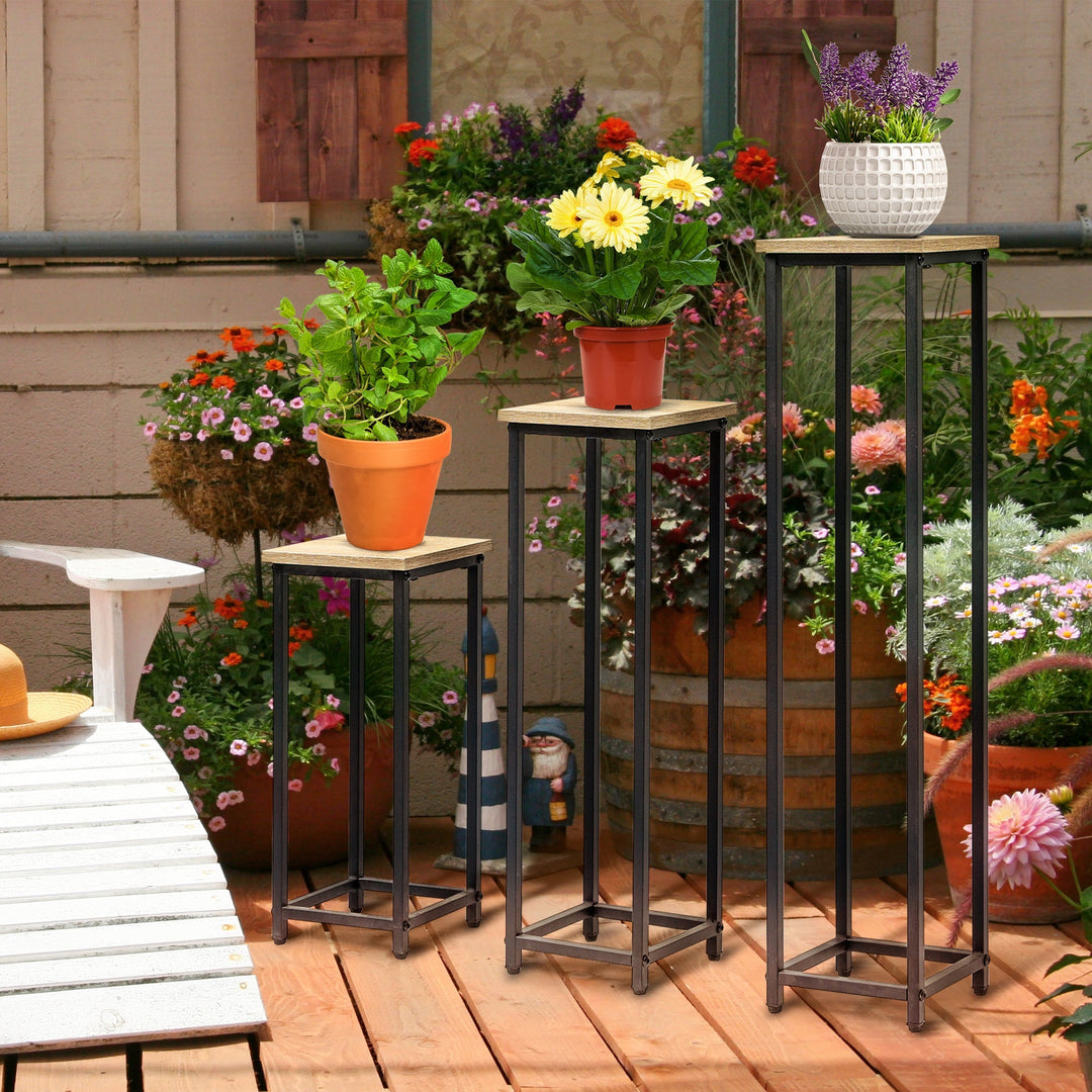 Tre plantestativer i metal og træ med potteplanter på en solrig træterrasse med blomster